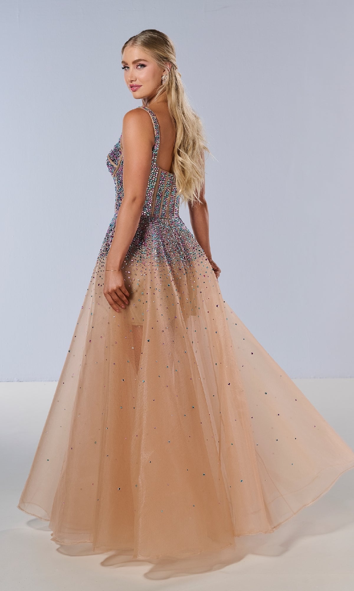 A woman poses in the Tiffany 16206 Long Prom Dress, a sleeveless, floor-length tulle gown with a crystal-embellished corset bodice and a flowing sheer peach skirt dotted with delicate sparkles.